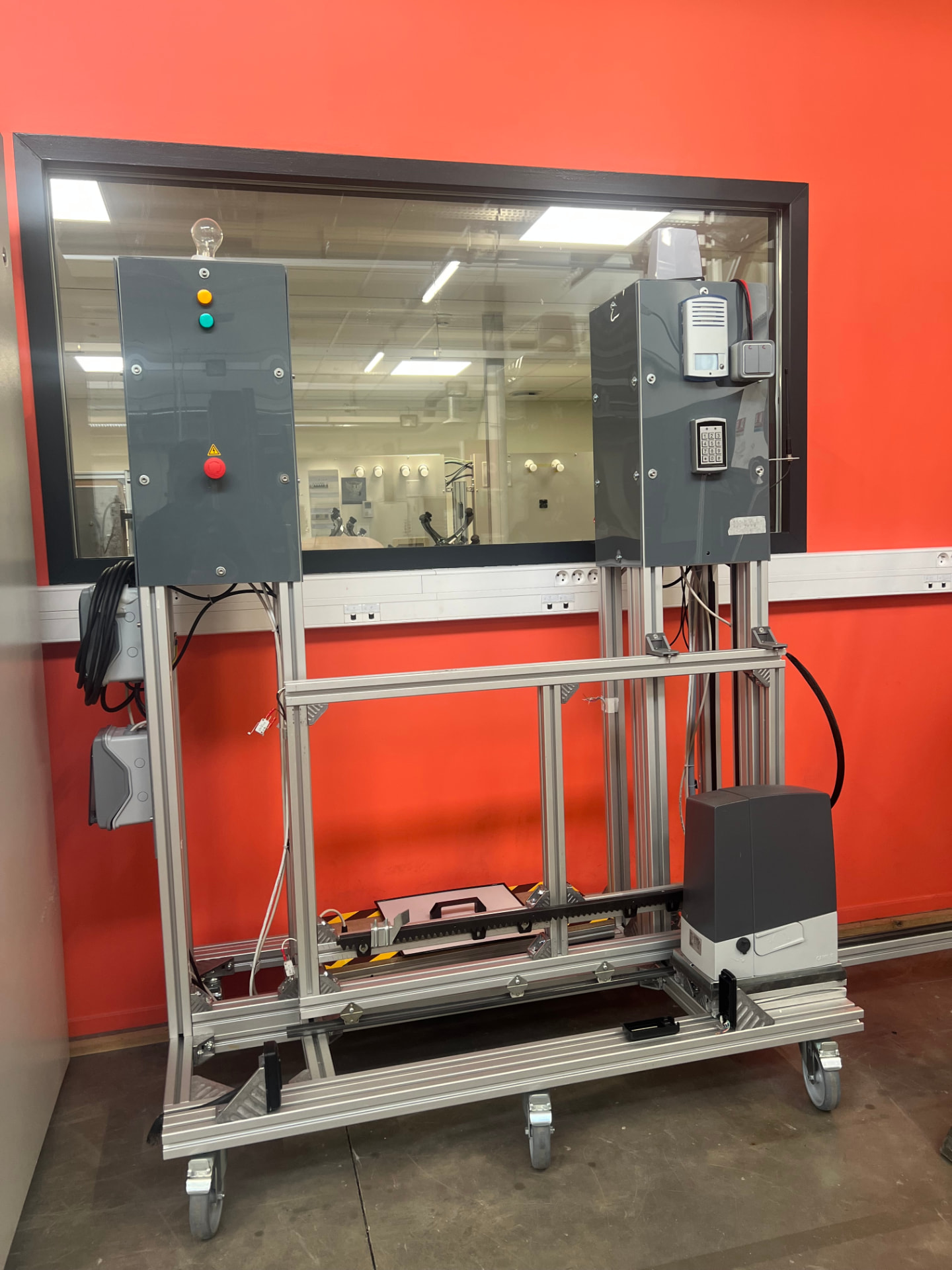 Banc d'essai pédagogique simulant un portail coulissant motorisé : structure en profilés d'aluminium sur roulettes, armoire de commande avec voyants et bouton d'arrêt d'urgence rouge, digicode et interphone, moteur électrique et crémaillère dentée, bandes de sécurité jaune et noir, devant un mur rouge-orangé.